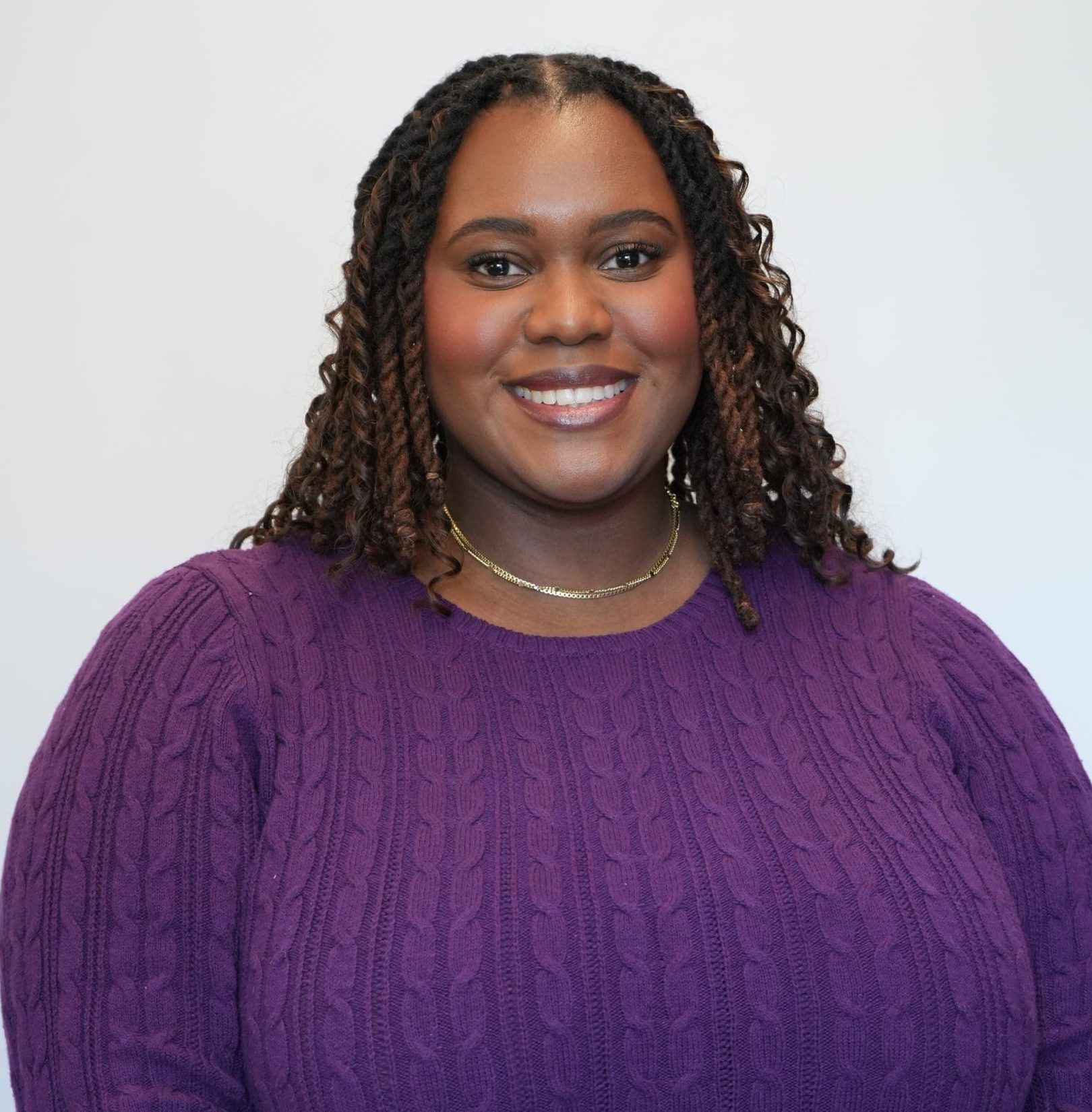 Portrait of a smiling woman with braided hair wearing a purple sweater against a light gray background, facing the camera.